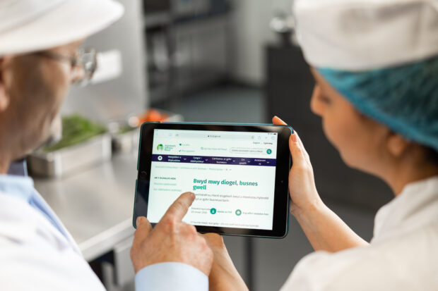 An environmental health officer shows a chef in a hairnet the FSA website on a tablet computer. We're looking over they're shoulders at the tablet screen.