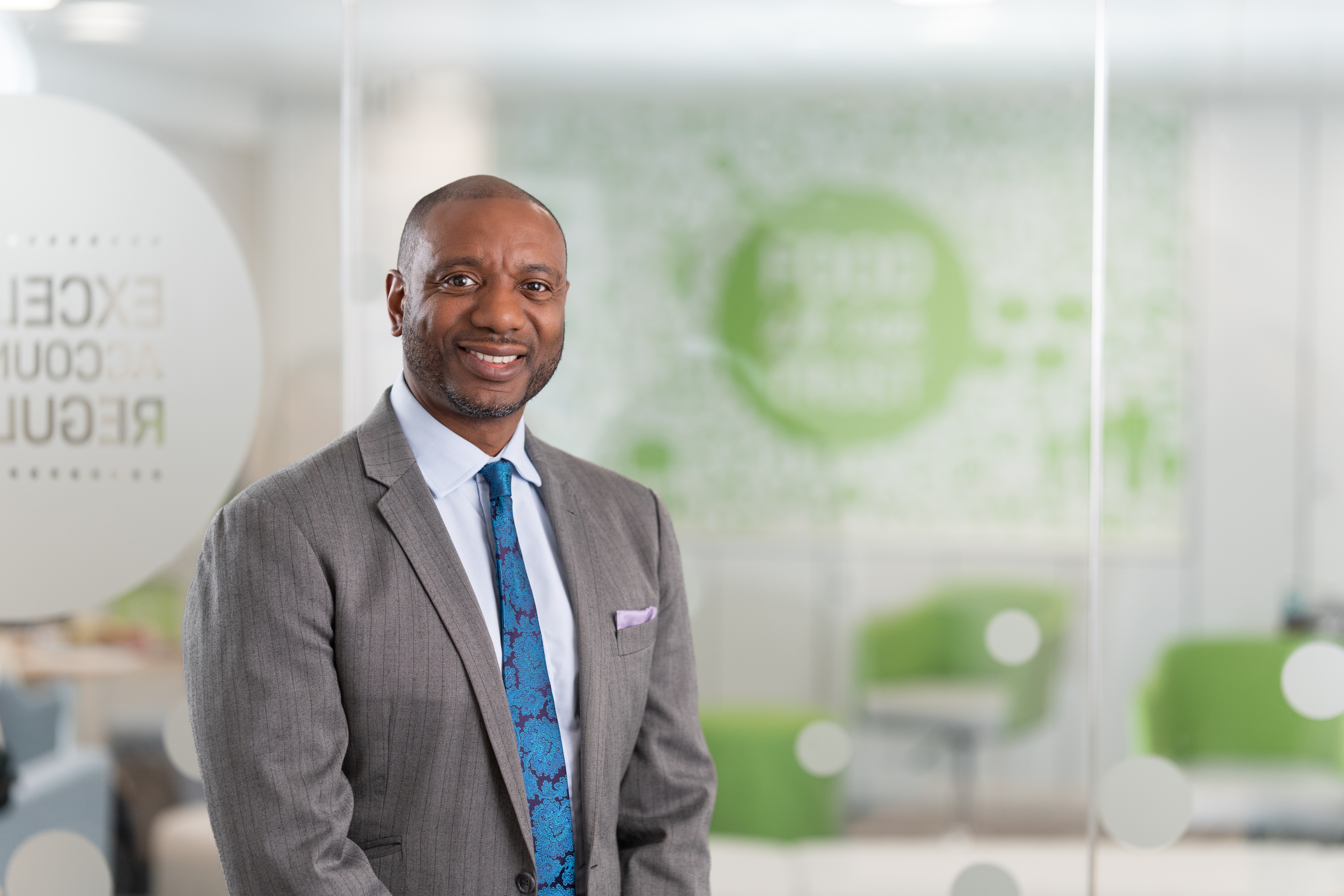 A man wearing a grey textured suit jacket, a white shirt, and a blue patterned tie stands in front of glass panels in a modern office.