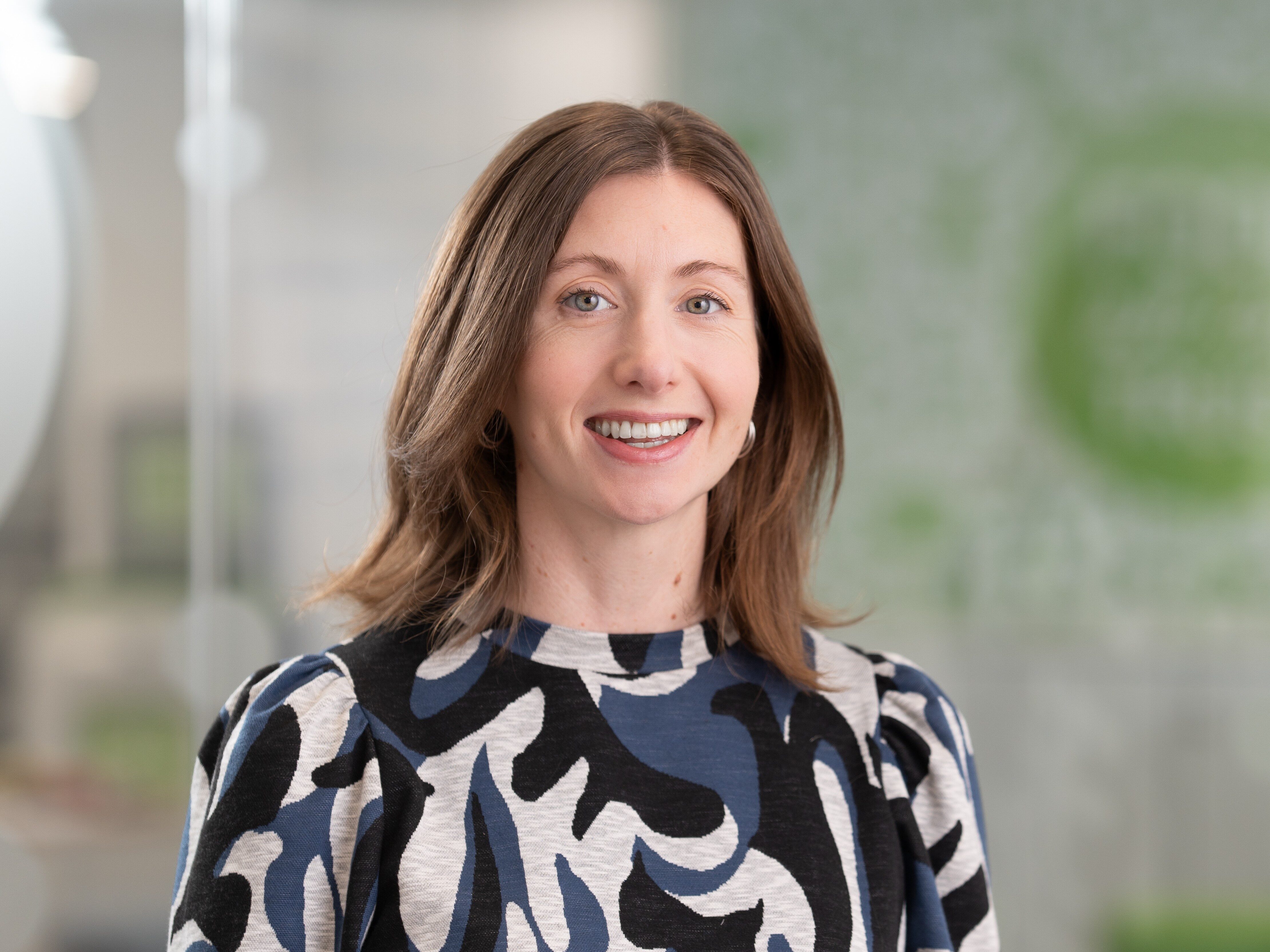 A woman with shoulder length brown hair, wearing a patterned top, in an office setting.